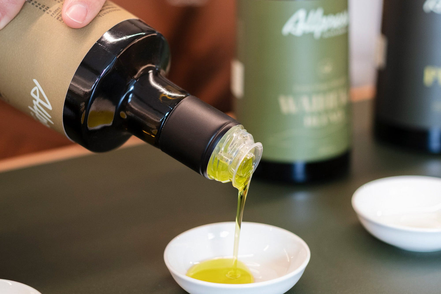 Allpress Olive Grove Olive Oil tasting Room. Bottle of olive oil being poured into a small white bowl with other bottles in the background.