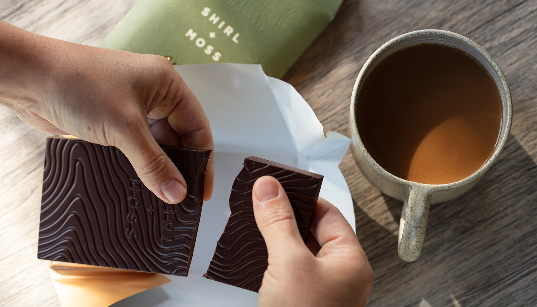 Olive Oil Chocolate Oil Shirl + Moss. Hands holding chocolate bars with a mug of coffee on a wooden surface